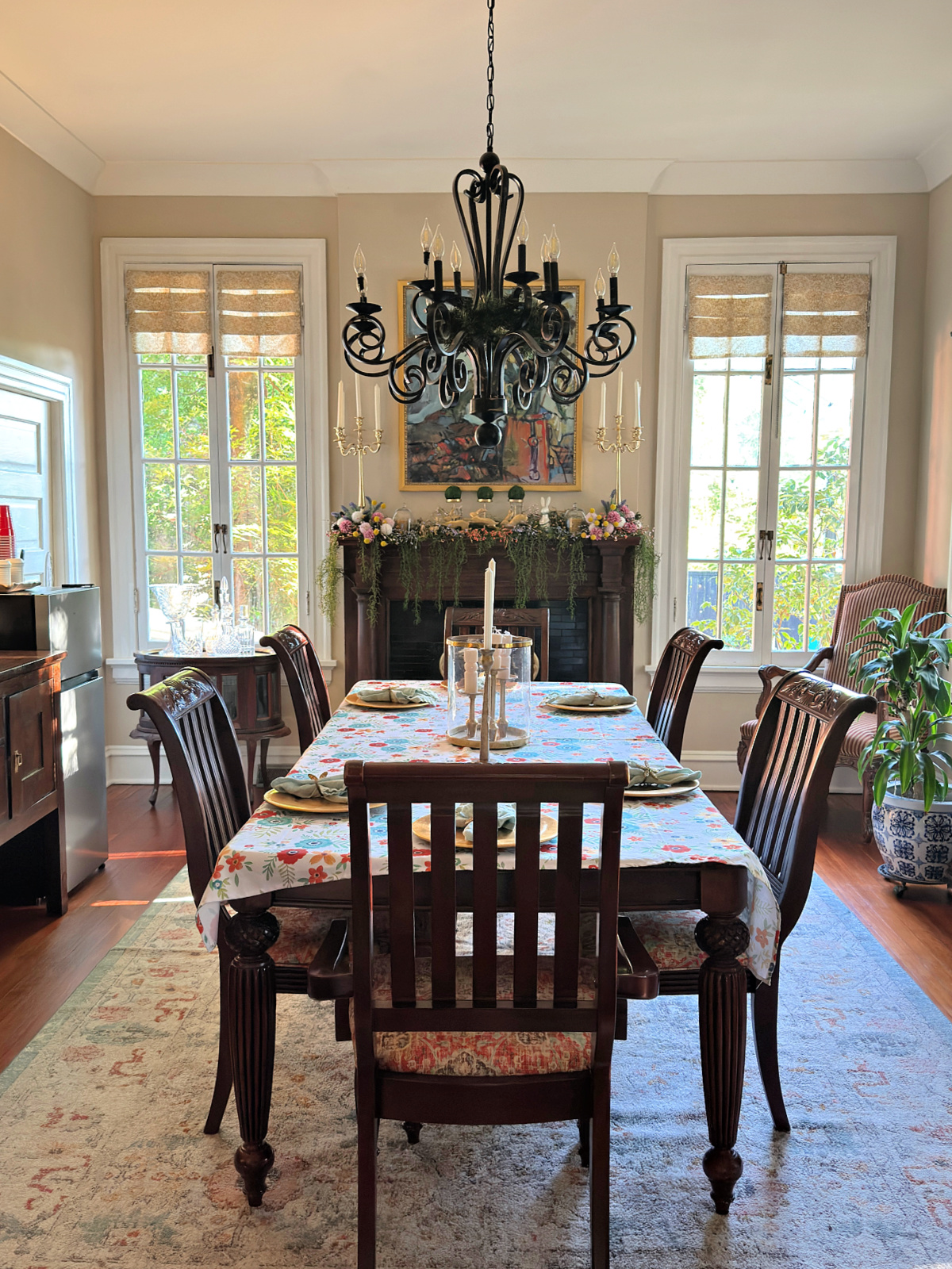 Elegant dining room with chandelier, set table, fireplace mantel decorated with florals, and tall windows letting in natural light.