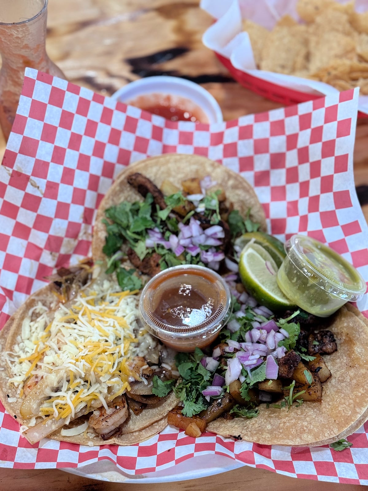 Three street-style tacos topped with cilantro and red onion, served with lime wedges and salsa on red checkered paper.