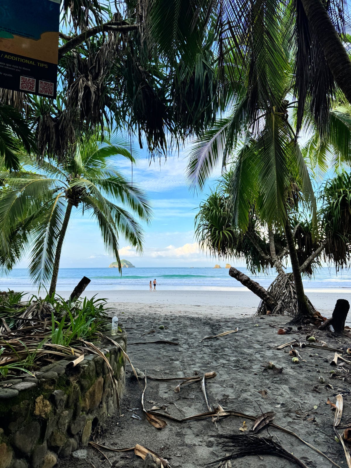 View of Manuel Antonio beach framed by palm trees, with two people walking along the sandy shore and small islands visible offshore.