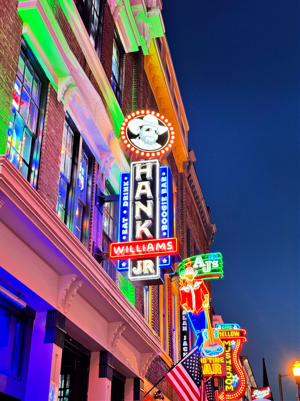 Neon bar signs light up historic brick buildings along Broadway in downtown Nashville at dusk.