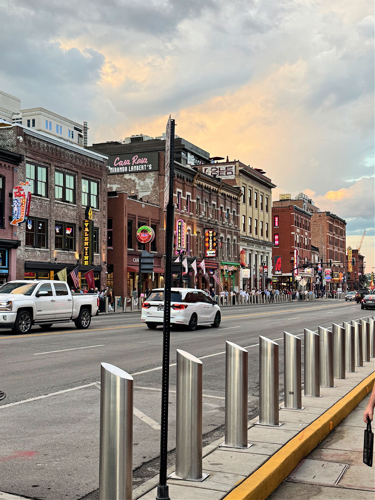 Honky-tonks and neon signs line Broadway in downtown Nashville at sunset with traffic and pedestrians.