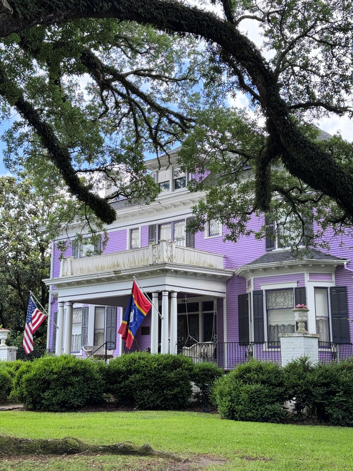 Large historic purple home with white columns, black shutters, and American flag beneath sprawling oak trees.