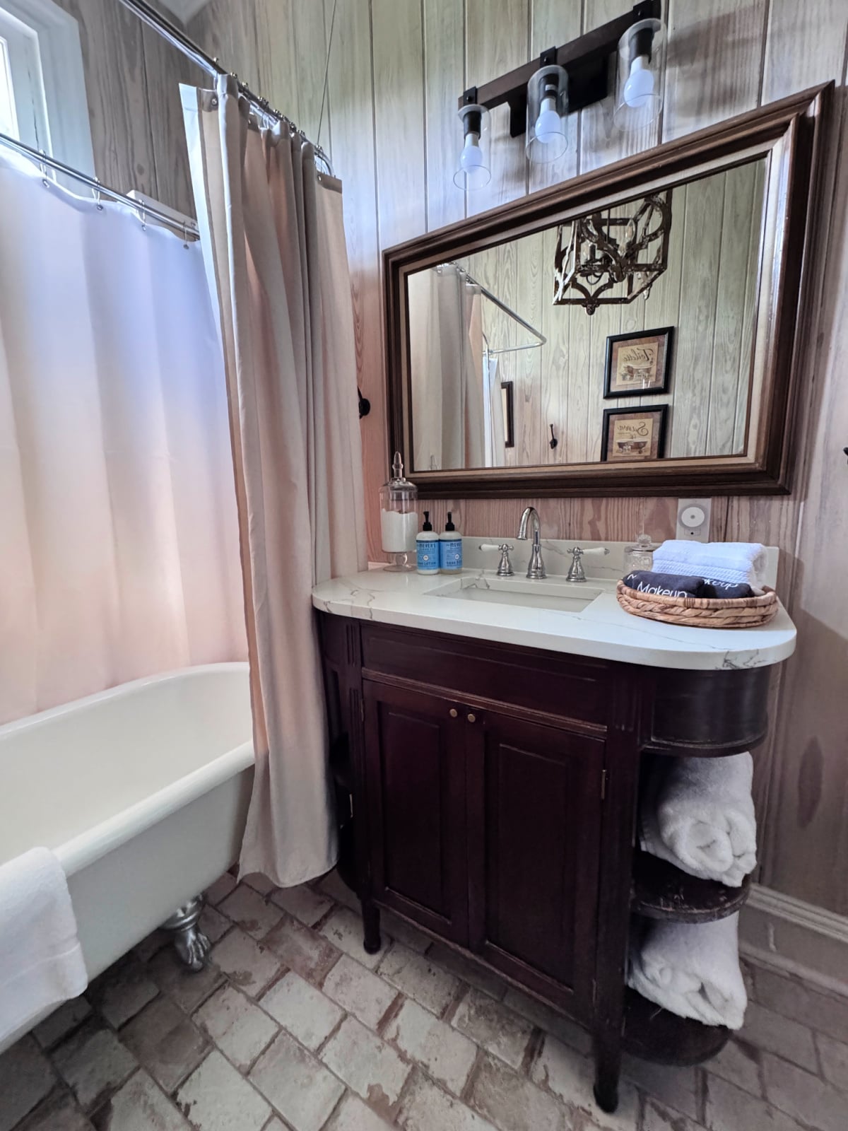 Vintage-style bathroom with clawfoot tub, wood vanity, large framed mirror, and brick flooring.