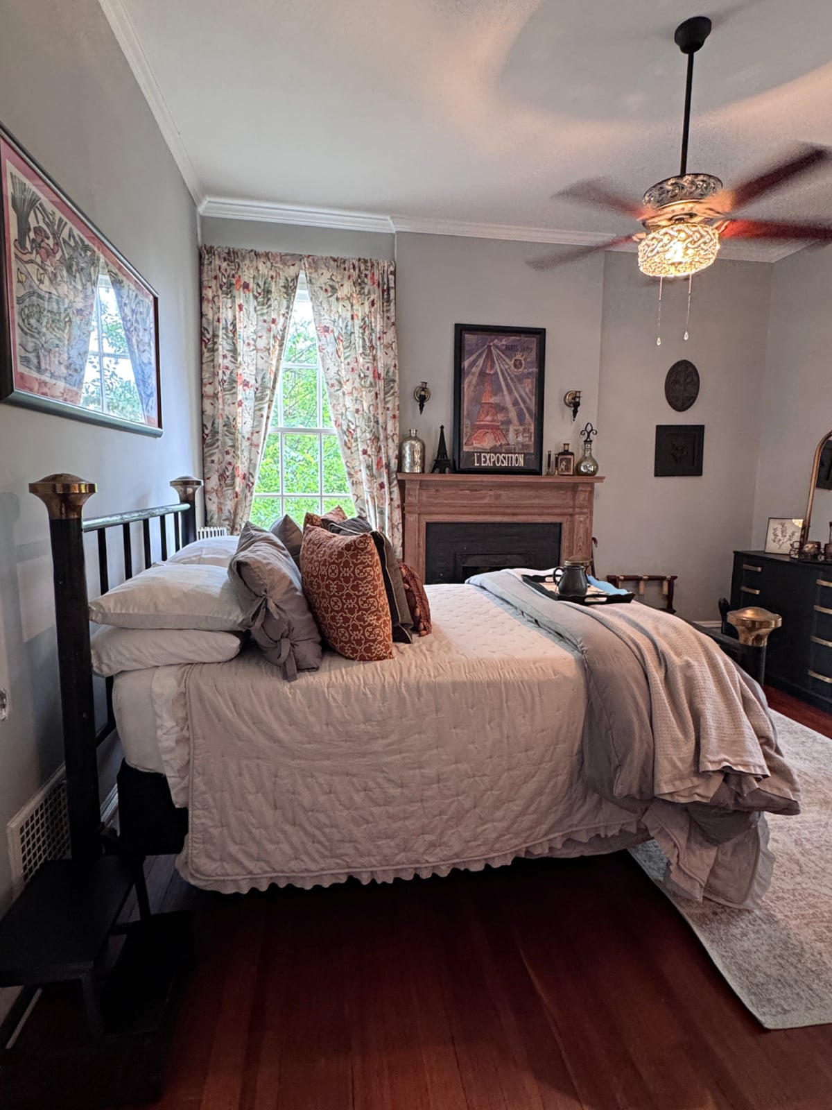 Cozy bedroom with iron bed frame, fireplace mantel, floral curtains, hardwood floors, and ceiling fan.