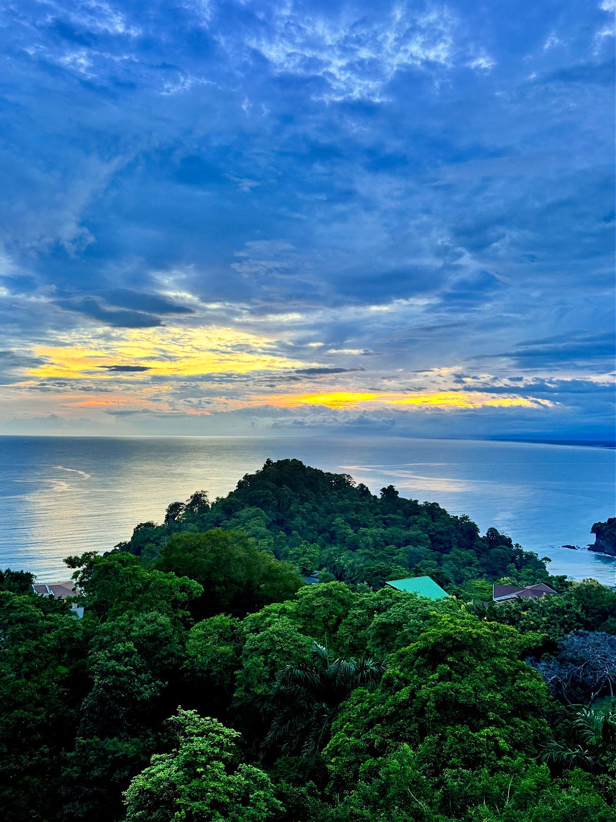 Colorful sunset over the Pacific Ocean in Manuel Antonio, Costa Rica, with golden light breaking through dramatic blue clouds above lush green hills.