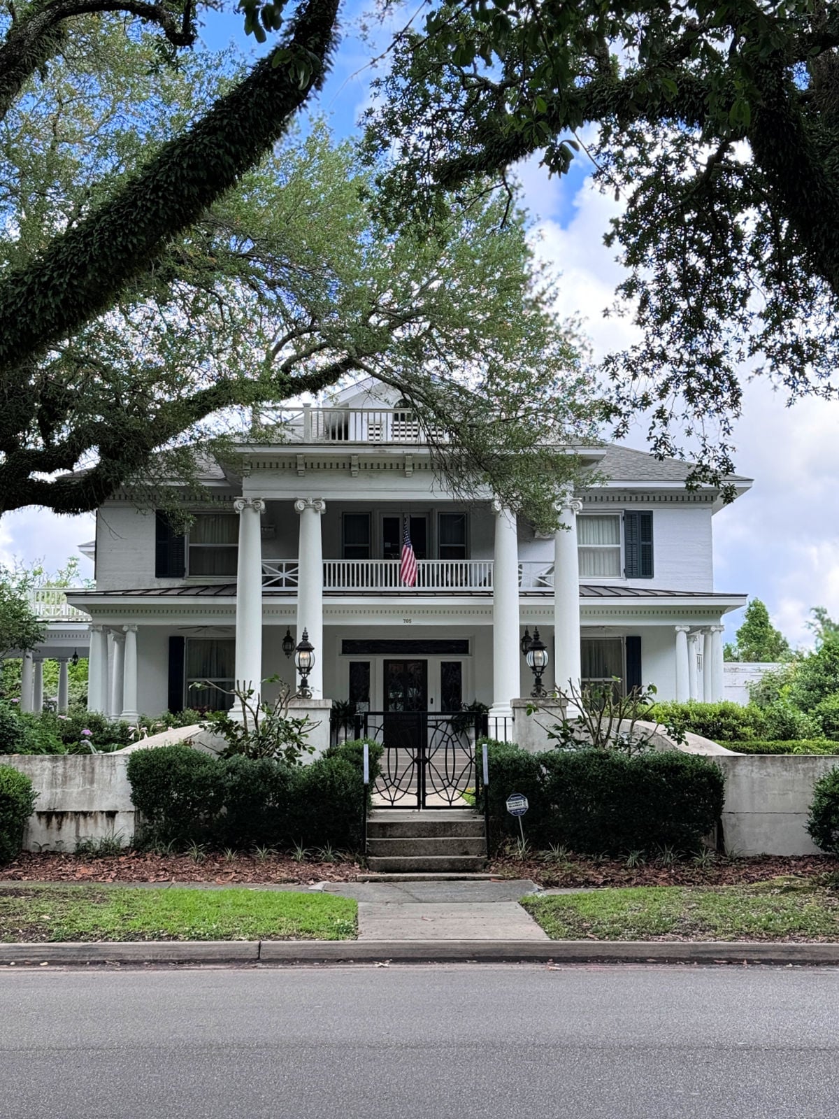 Large white colonial-style home with tall columns, balcony, and oak trees lining the front yard.