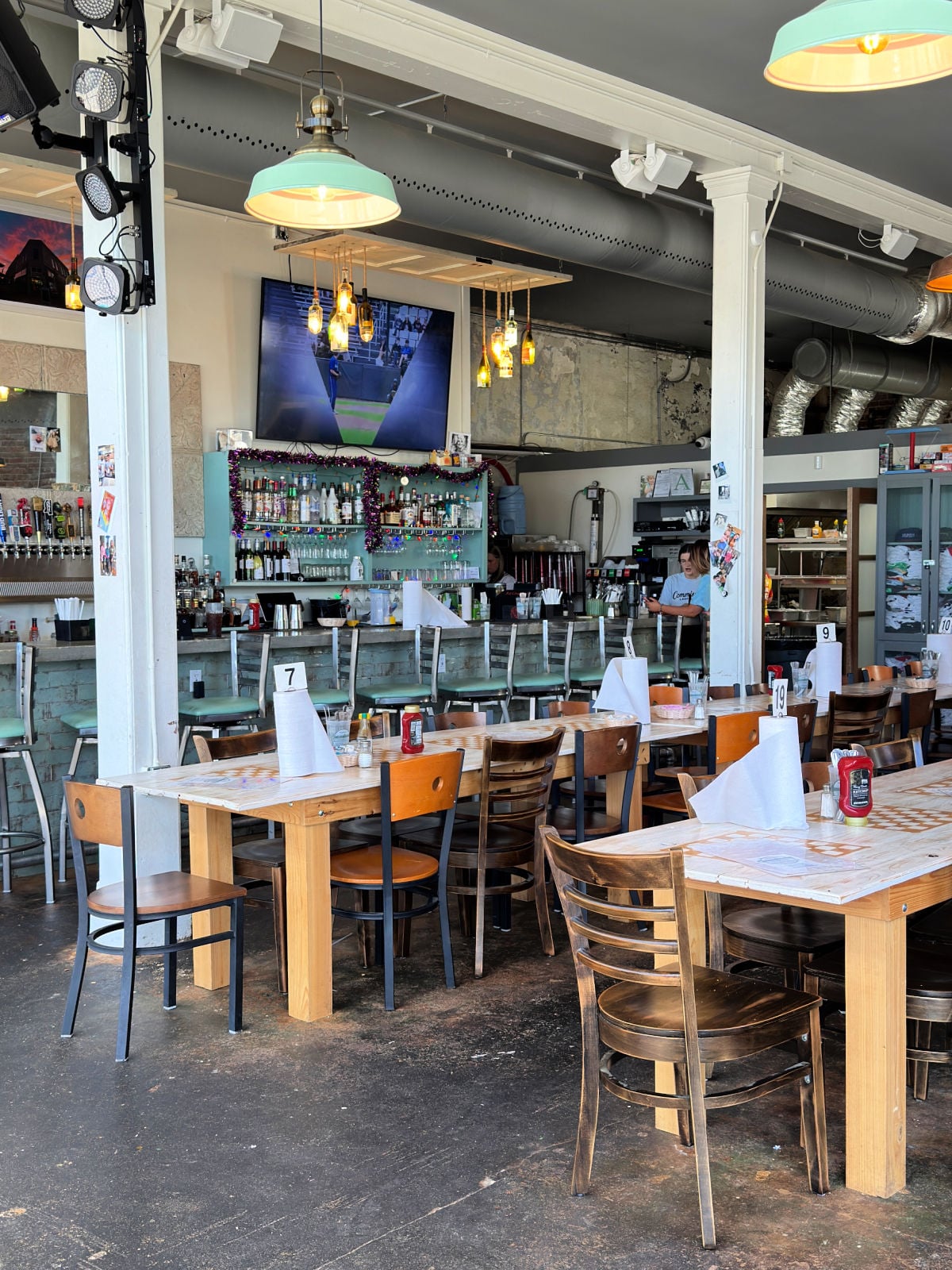 Industrial-style restaurant interior with wooden tables, bar seating, pendant lighting, and open kitchen.