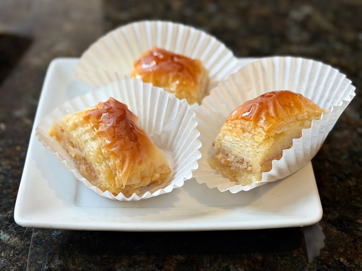 Baklava pastries in paper liners on a white plate, showing flaky layers and honey syrup.