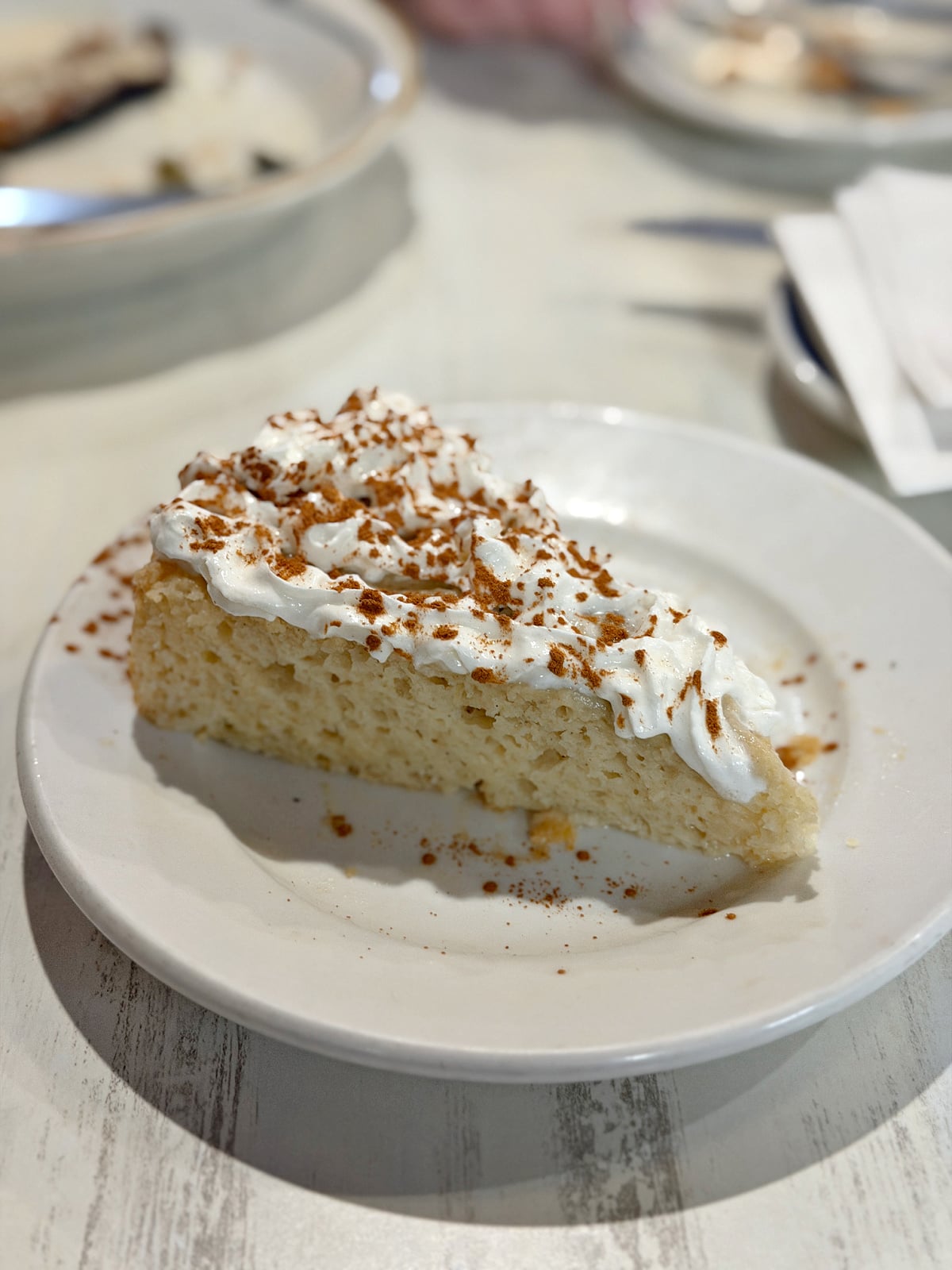 Slice of cake topped with whipped cream and cinnamon on a white plate at a restaurant.