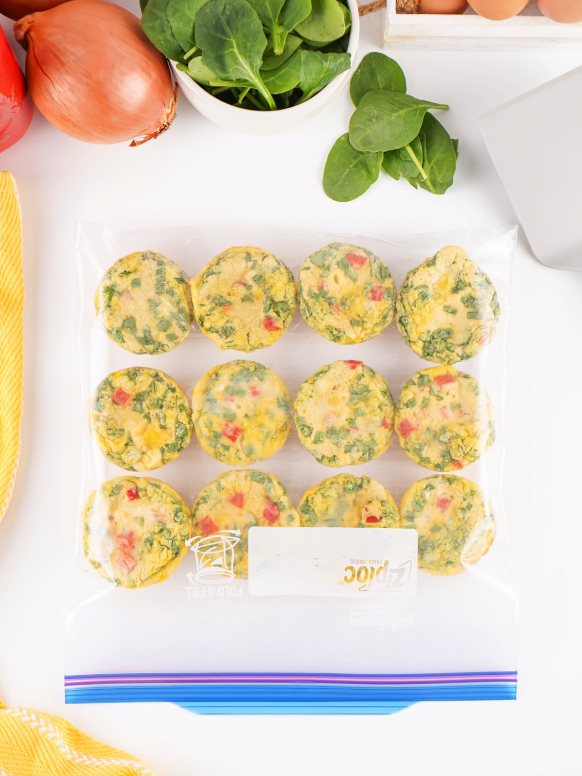 Freezer-friendly egg bites arranged in a zip-top freezer bag with fresh spinach, onion, eggs, and red bell pepper in the background.