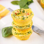 Stack of homemade egg bites filled with spinach, red bell pepper, and cheese on a white plate with fresh spinach garnish.