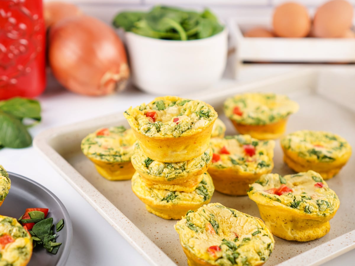 Homemade egg bites stacked on a tray with more egg bites surrounding them, with fresh spinach, eggs, onion, and red bell pepper in the background.