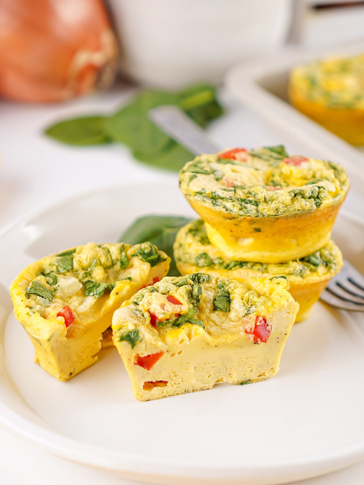 Egg bites on a white plate, including one cut open to show the fluffy interior with spinach, red bell pepper, and cheese.