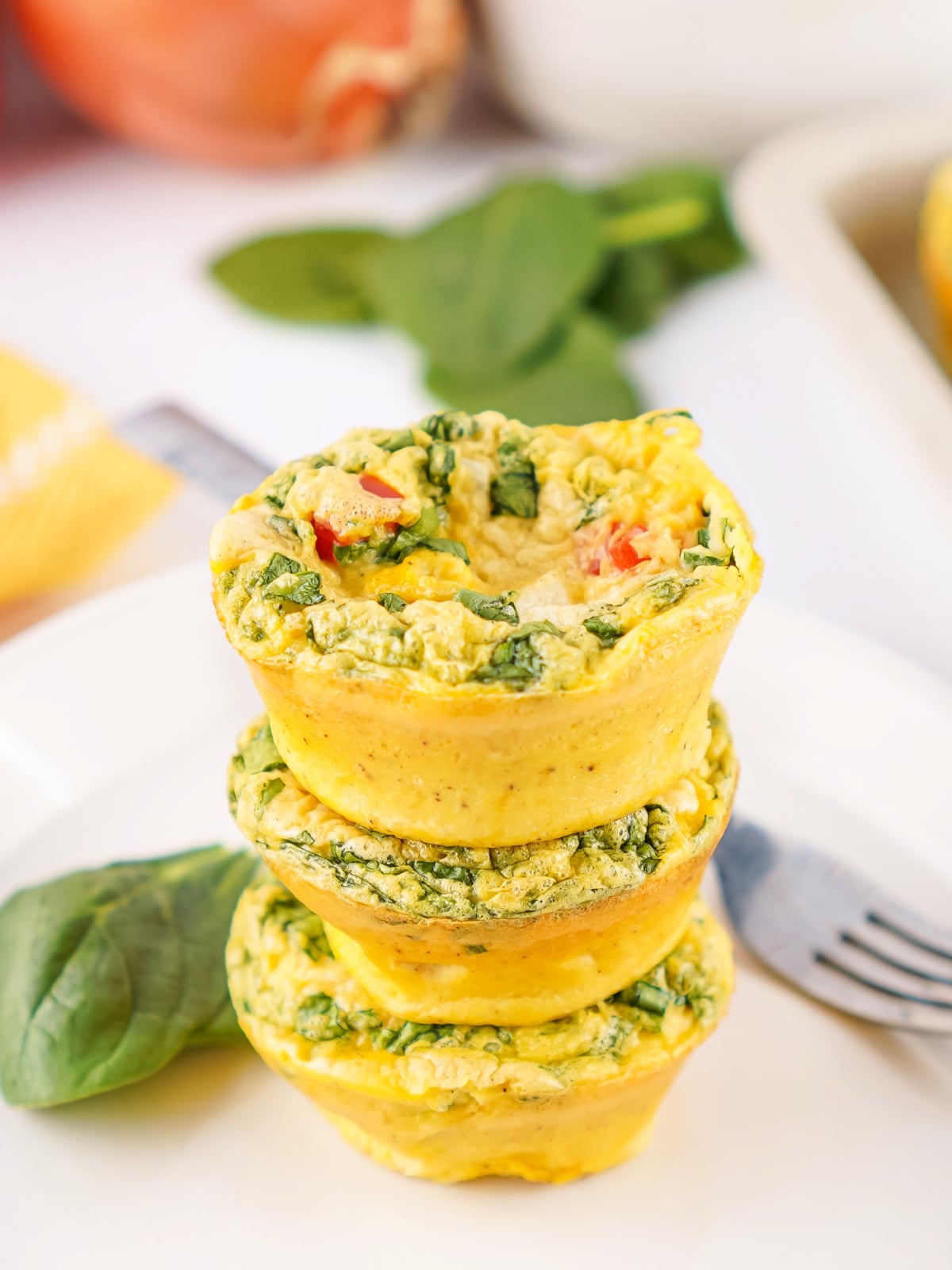 Three homemade egg bites stacked on a plate, filled with spinach, red bell pepper, and cheese, with a fork and fresh spinach nearby.