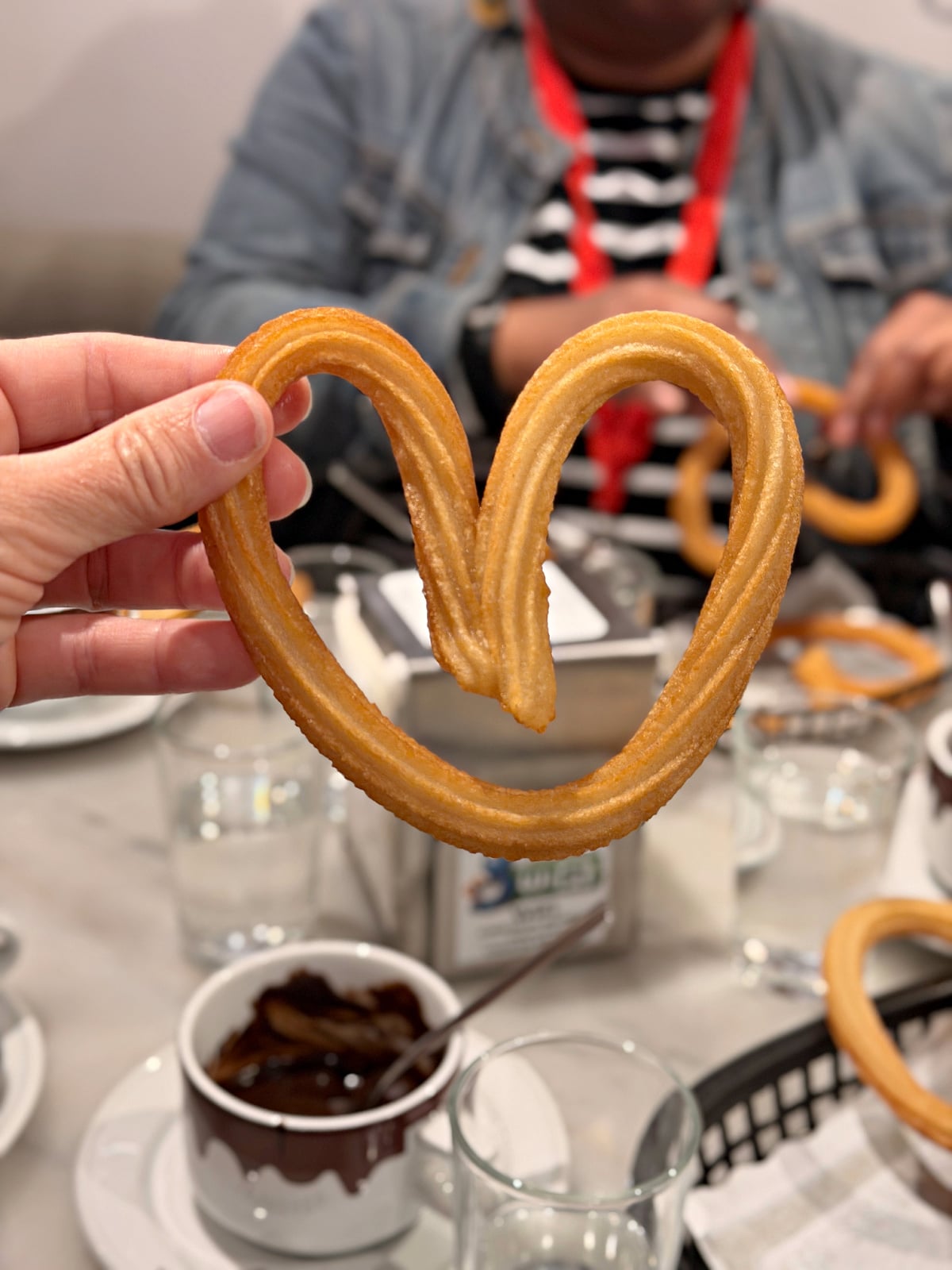 Heart-shaped churro held in front of cups of rich Spanish chocolate during breakfast in Madrid.