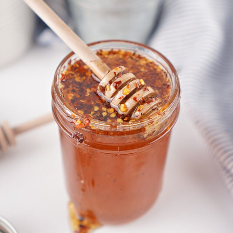 Close-up of a jar of homemade hot honey with a honey dipper resting on top and red pepper flakes visible throughout.