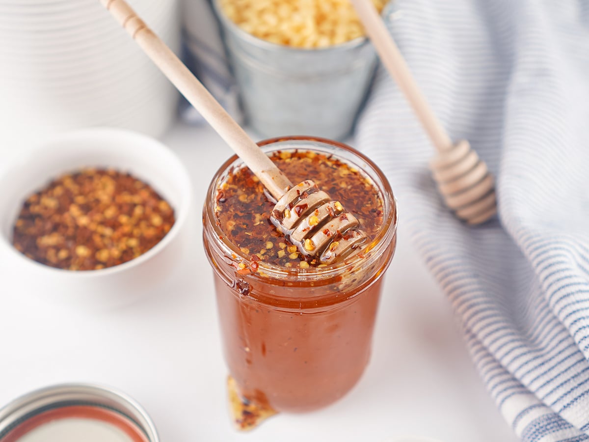 Jar of homemade hot honey topped with a honey dipper, with bowls of chili flakes and honey in the background.
