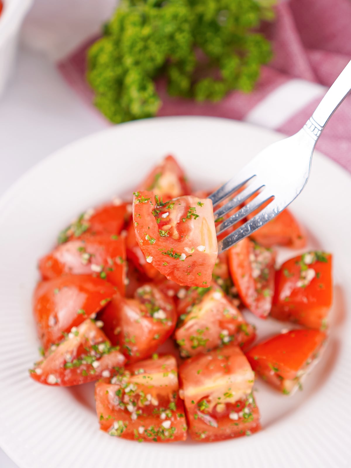 Fork lifting a juicy bite of marinated tomatoes coated in a fresh herb and garlic vinaigrette from a white plate.