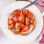 Fresh marinated tomatoes tossed in a garlic herb dressing served on a white plate with parsley and a striped kitchen towel.