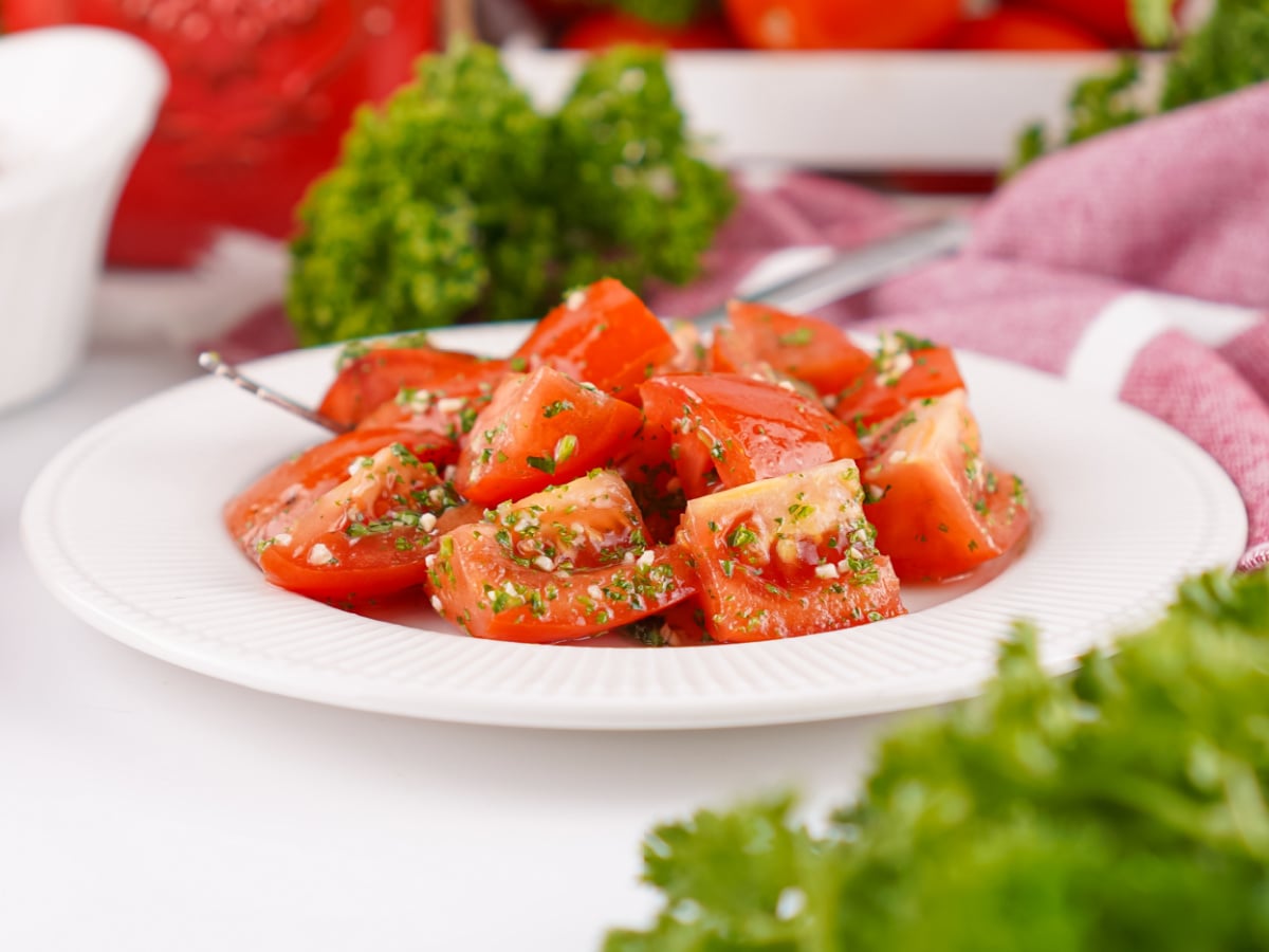 Plate of marinated tomatoes coated in a light herb vinaigrette, photographed from the side with fresh parsley in the foreground.