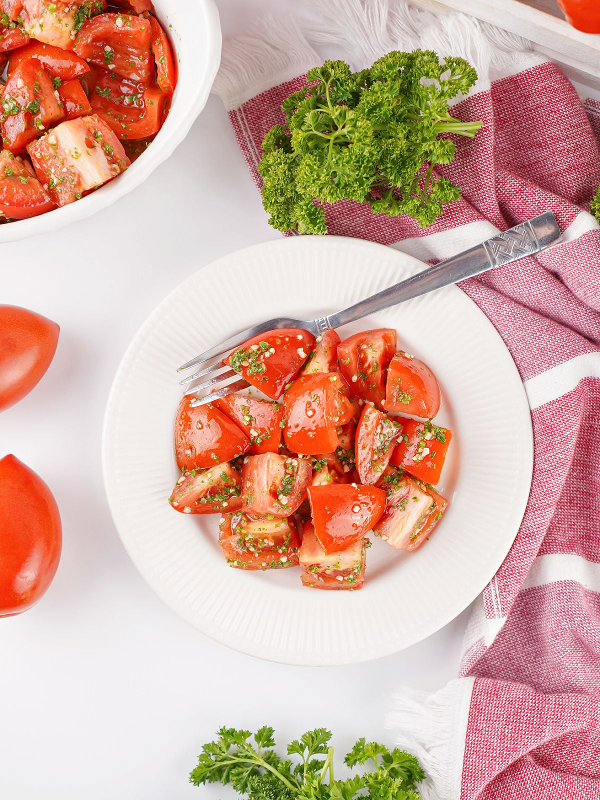 Marinated tomato salad on a white plate with a fork, fresh parsley, and extra tomatoes styled around the dish for serving.