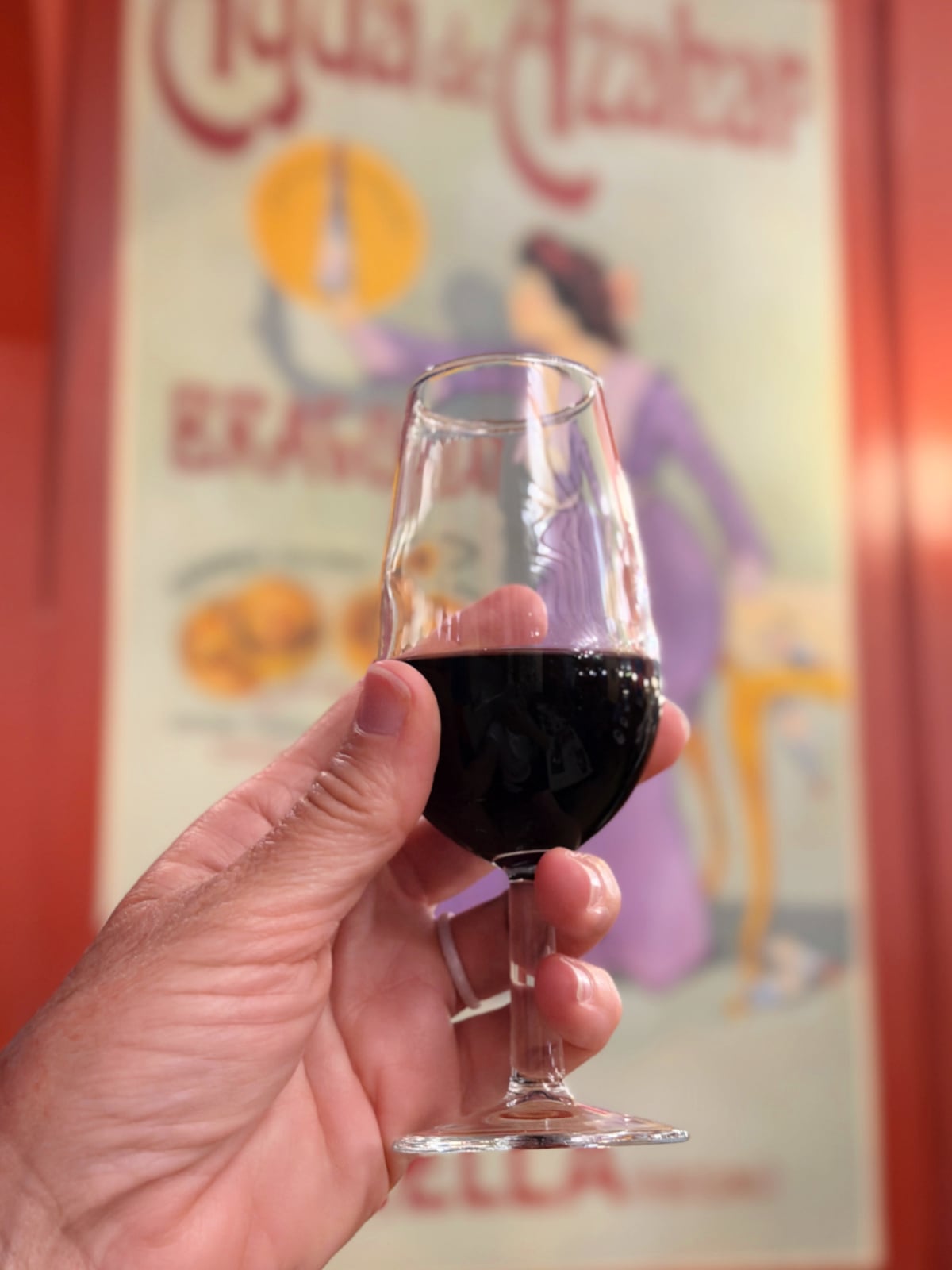 Small glass of Spanish red wine held in front of a vintage Madrid café poster.
