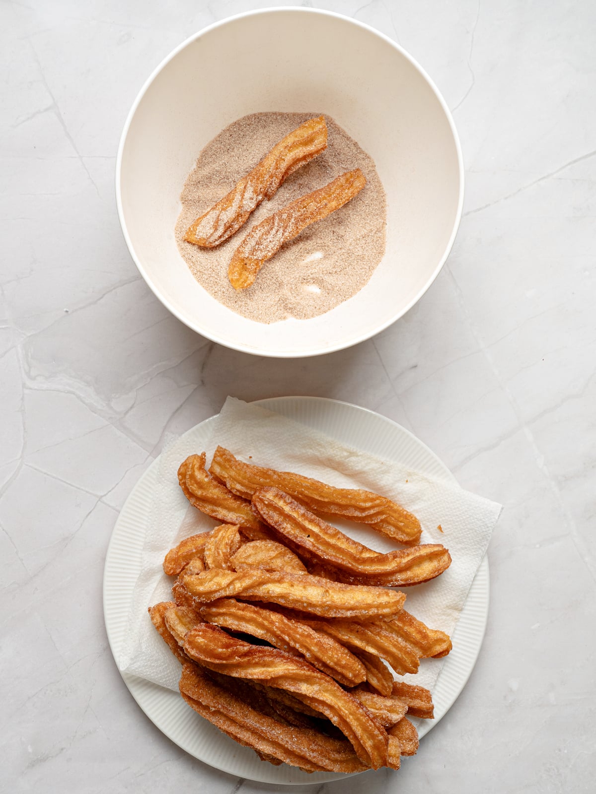 Freshly fried churros being coated in cinnamon sugar in a large white bowl.