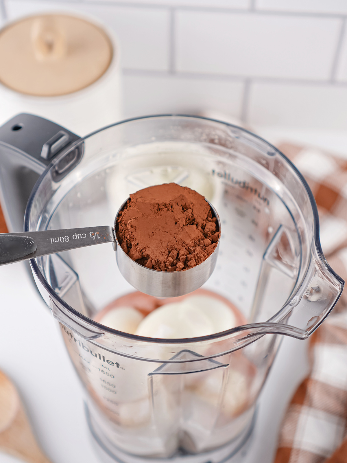 Measuring cup of cocoa powder being added to a blender with peeled hard-boiled eggs and milk for chocolate pudding.