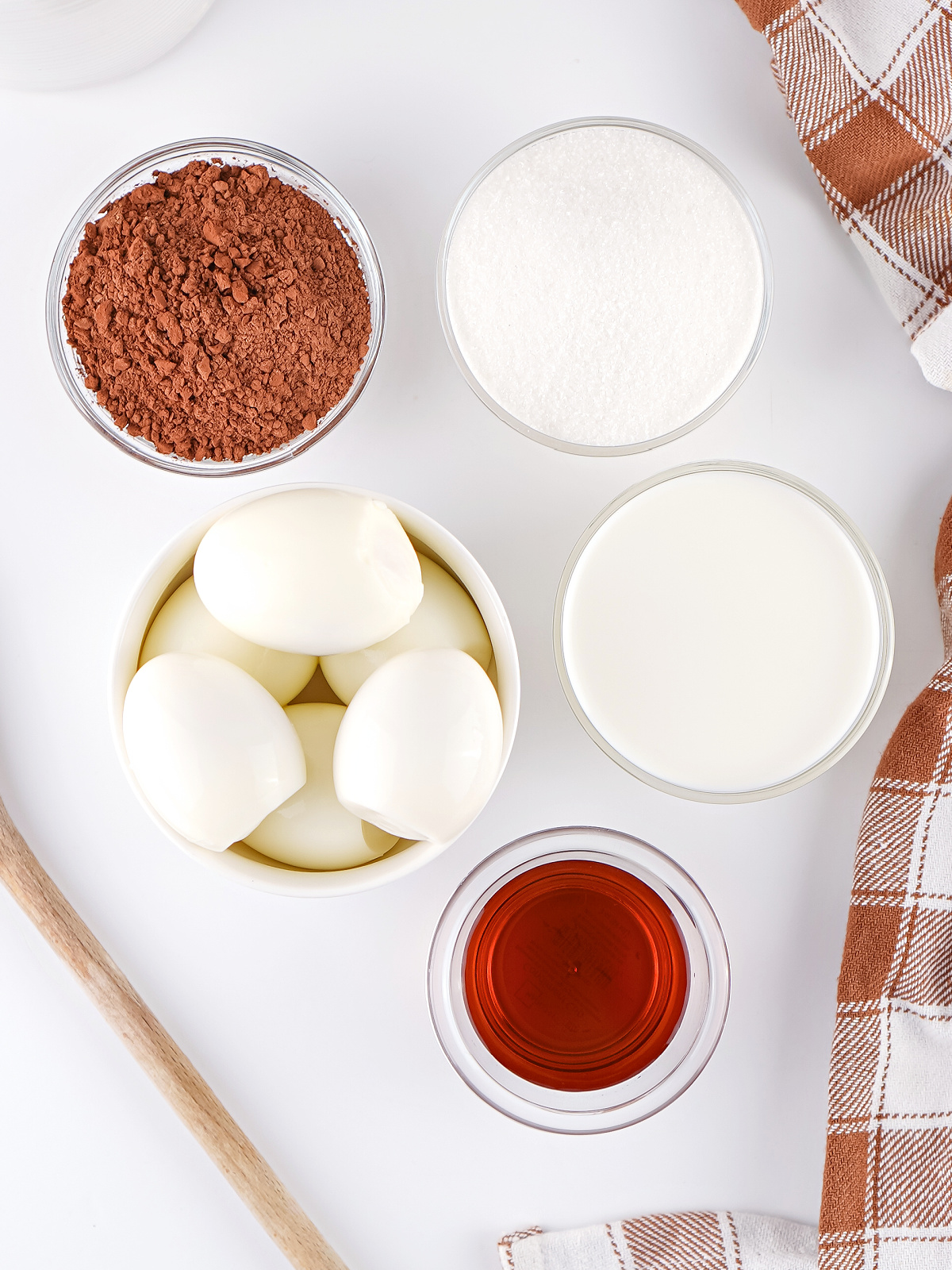 Ingredients for hard-boiled egg pudding including peeled eggs, cocoa powder, sugar, milk, and vanilla with a wooden spoon nearby.