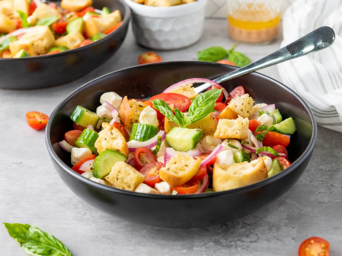 Panzanella salad with toasted ciabatta croutons, cherry tomatoes, cucumbers, mozzarella, red onion, and fresh basil in a black serving bowl.
