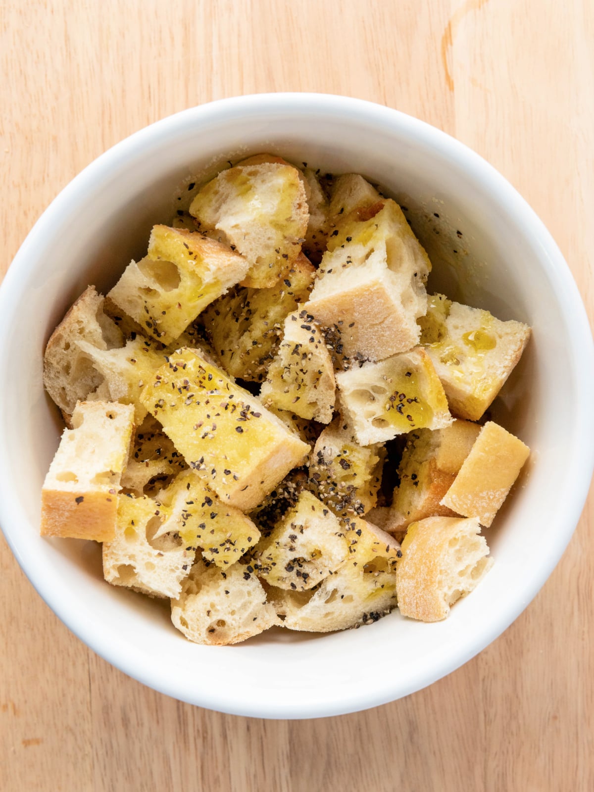 Cubed ciabatta bread tossed with olive oil and black pepper in a white bowl before toasting.