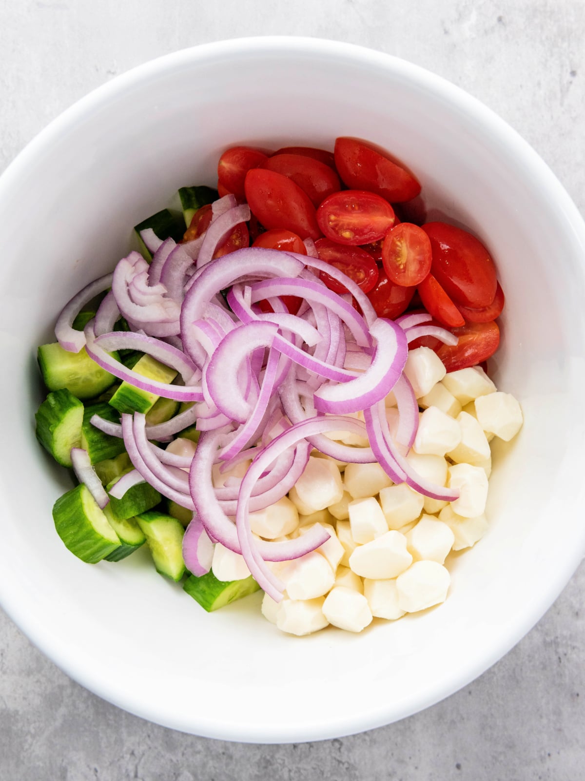 Cherry tomatoes, diced cucumbers, mozzarella pearls, and sliced red onion in a white mixing bowl for panzanella salad.