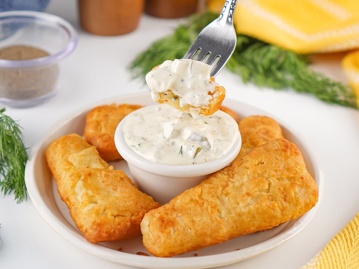 Crispy fish sticks arranged around a bowl of creamy homemade tartar sauce with a fork dipping in for a bite.