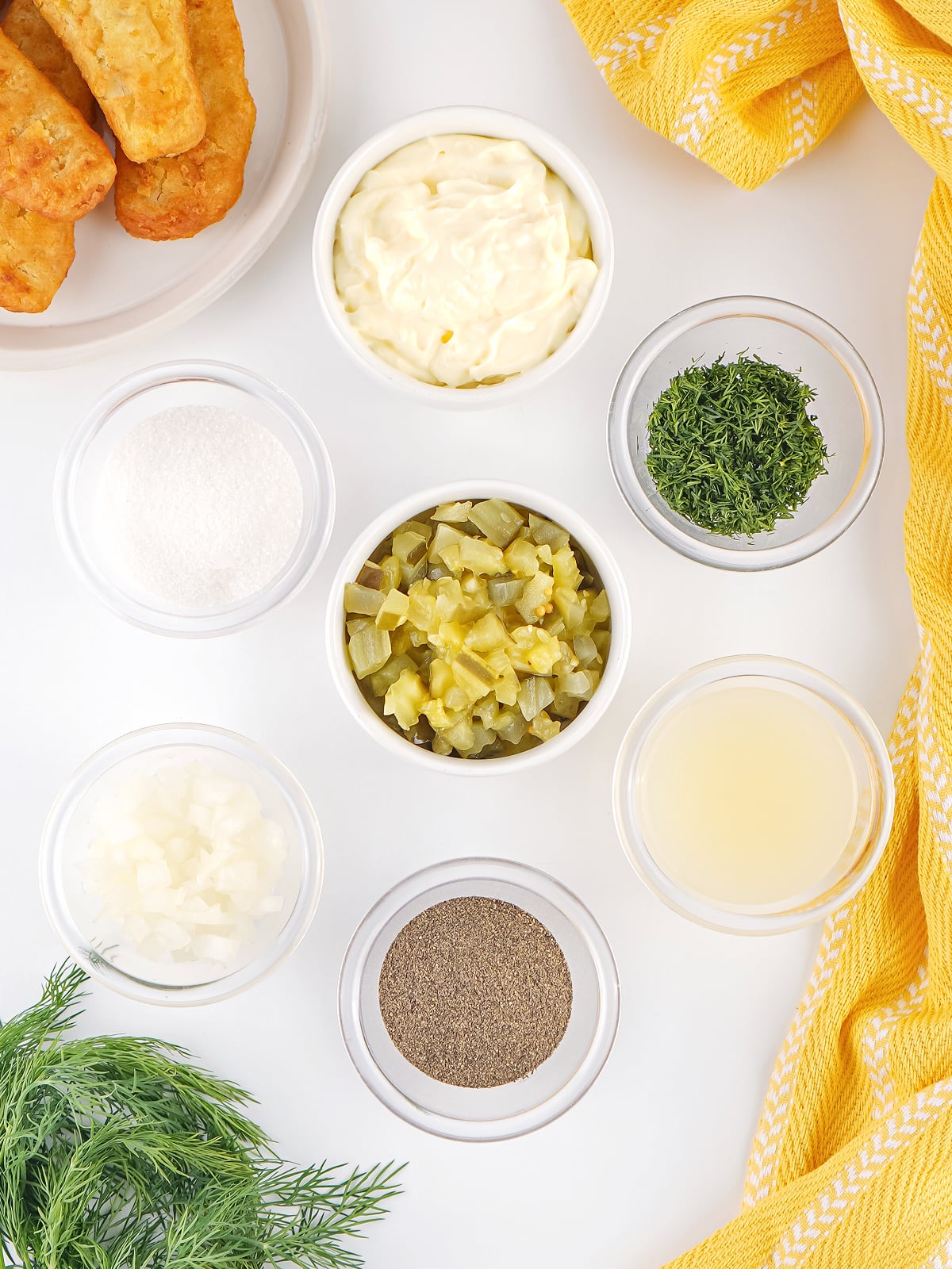 Ingredients for homemade tartar sauce arranged on a white surface, including mayonnaise, chopped pickles, onion, dill, lemon juice, salt, and pepper.
