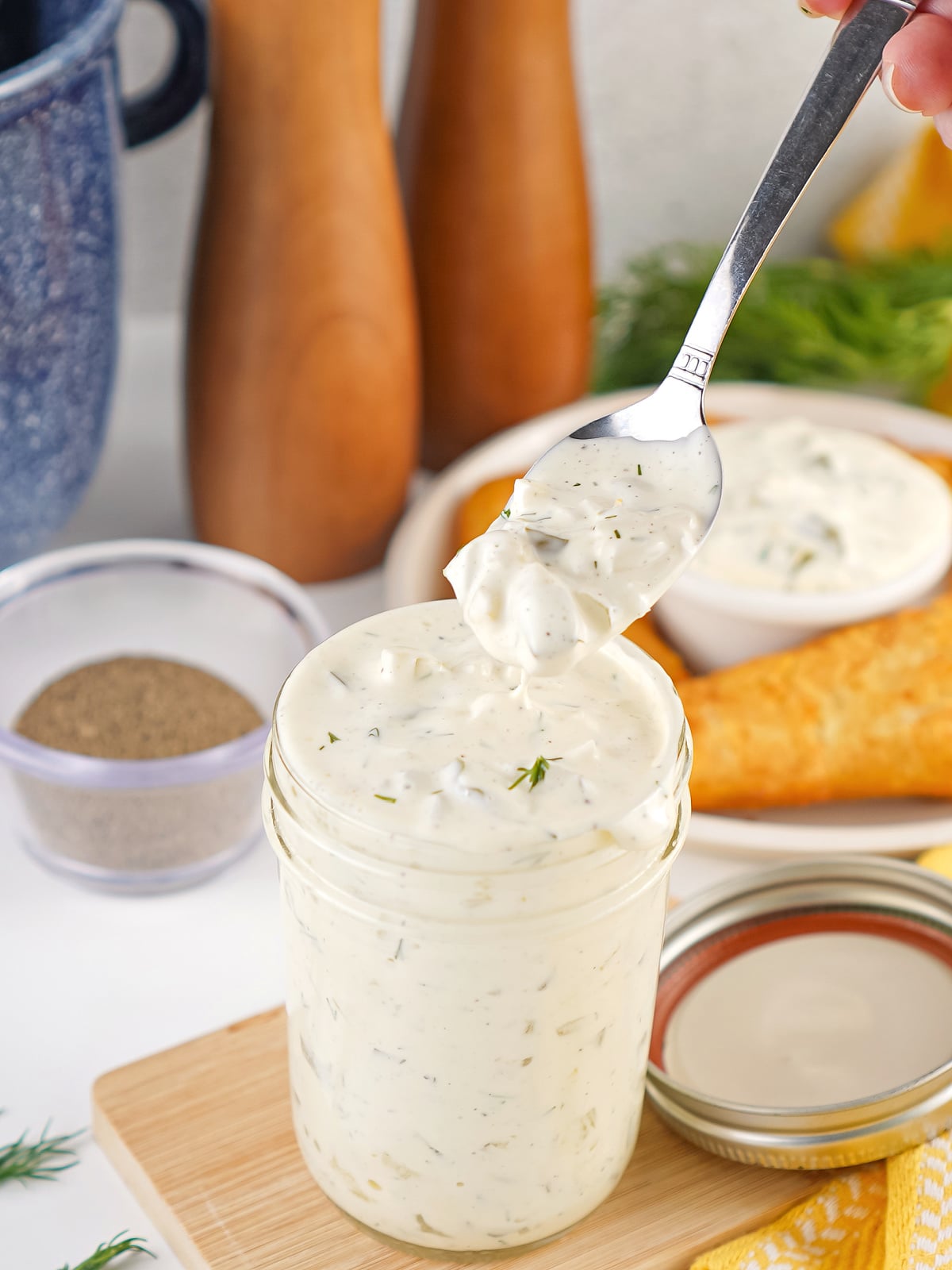 Spoon lifting a creamy scoop of homemade tartar sauce from a glass jar, showing the chunky texture with pickles and fresh dill.