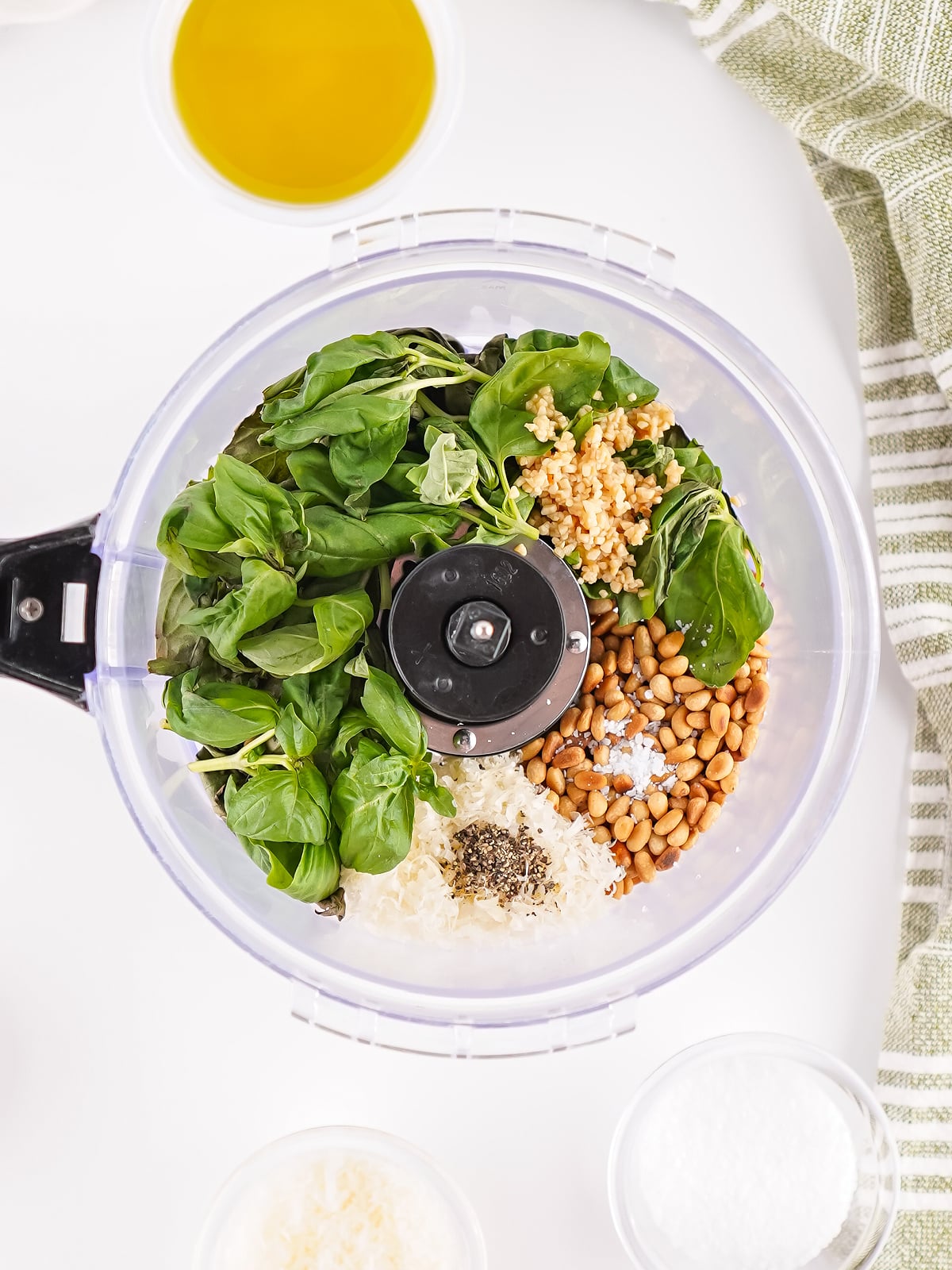 Fresh basil, toasted pine nuts, Parmesan cheese, garlic, olive oil, salt, and pepper in a food processor ready to blend into homemade pesto.