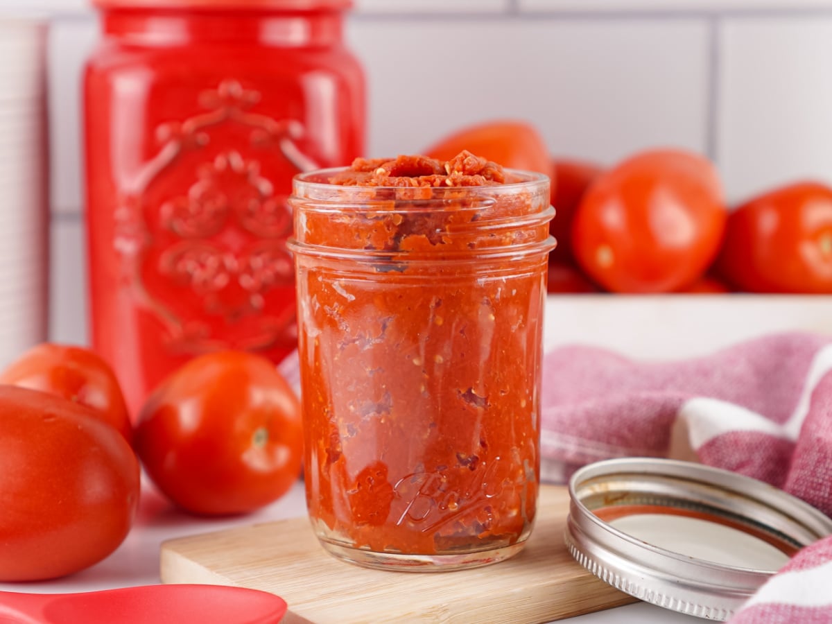 Homemade rustic tomato paste in a glass jar surrounded by fresh Roma tomatoes on a kitchen counter.