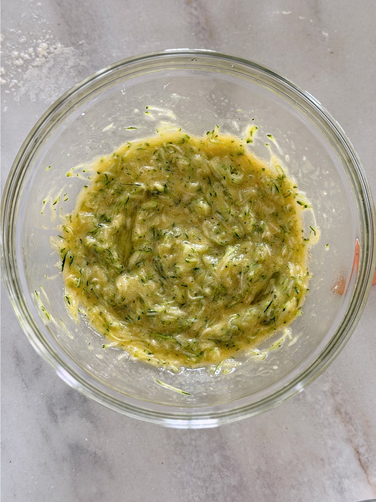 Shredded zucchini combined with the wet ingredients in a glass bowl.