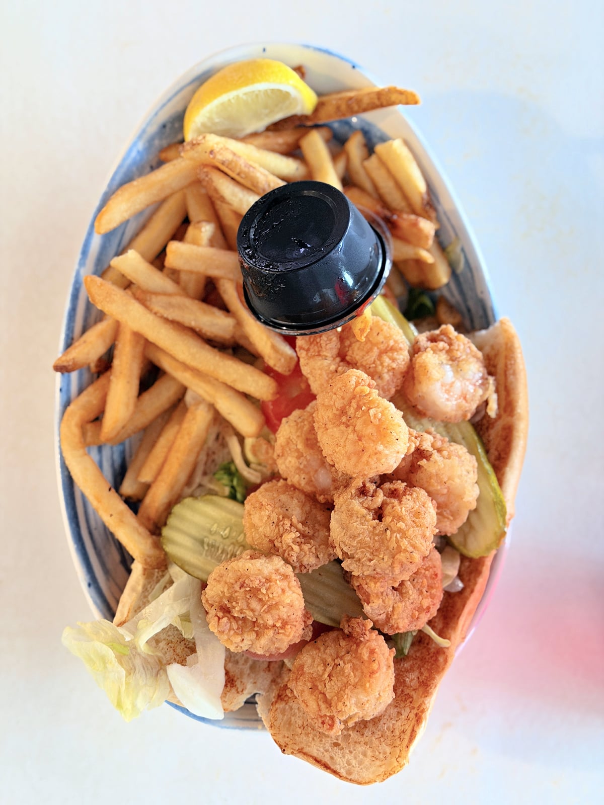 Fried shrimp Po’ Boy with lettuce, tomato, pickles, French fries, lemon, and dipping sauce.